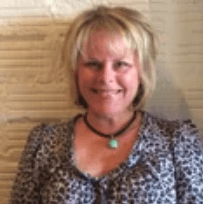 A smiling woman with short hair wearing a patterned blouse and a necklace against a wooden backdrop.